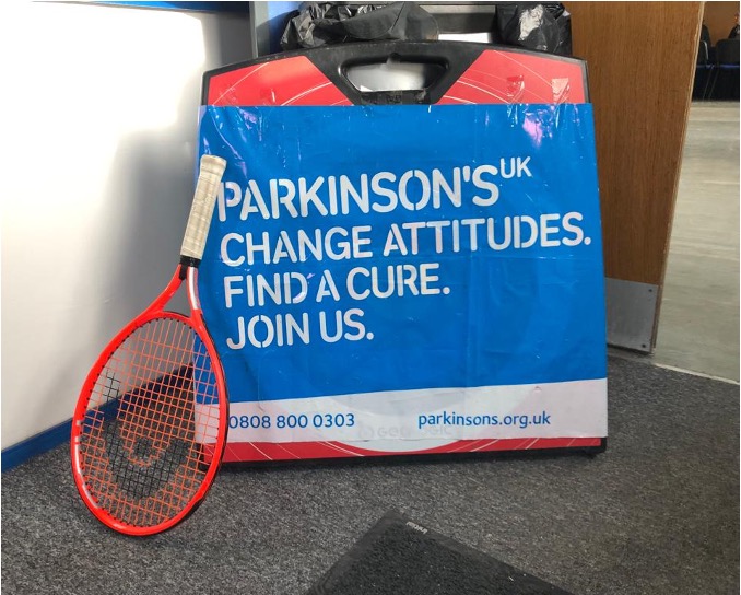 Tennis racket leaning against a blue poster sign which says: Parkinson's UK Change Attitudes. Find a cure. Join us